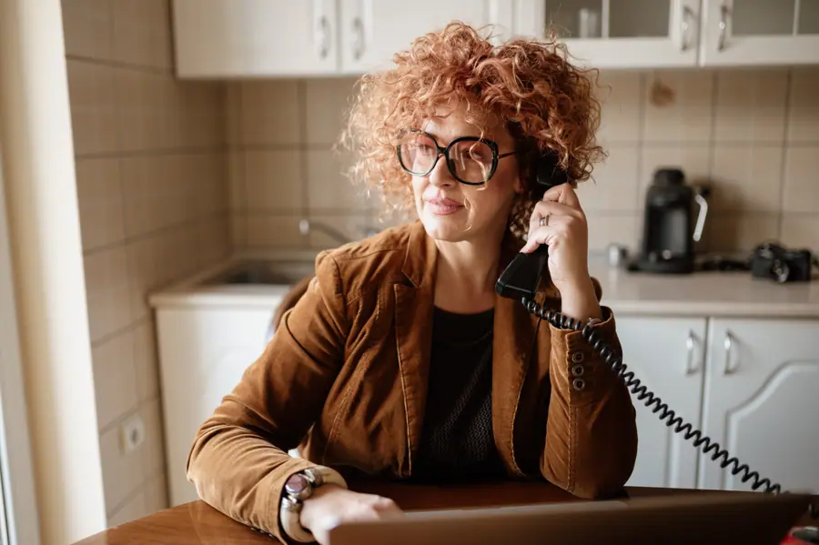 A woman with curly red hair interacts on a landline phone while seated at a kitchen table