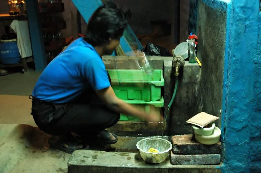 Washing glass mugs by hand in the basement interior of the chaang house, blue walls, blue shirt, motion, green boxes, soap, bricks, hose, spigot, Boudha, Kathmandu, Nepal (5894196563)