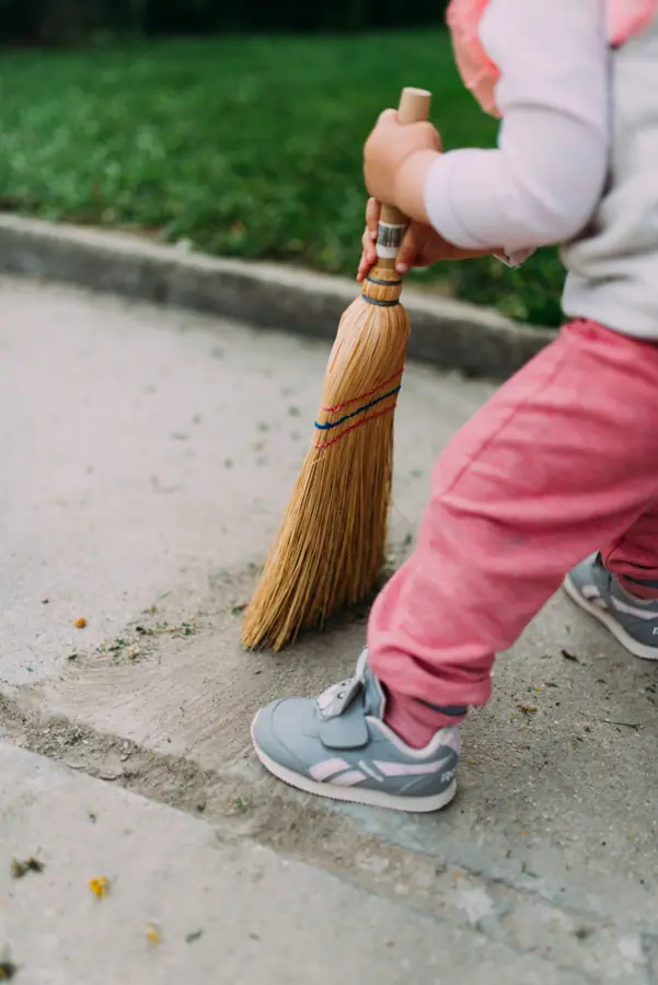 Child cleaning the ground with a broom in a grassy area during a sunny day