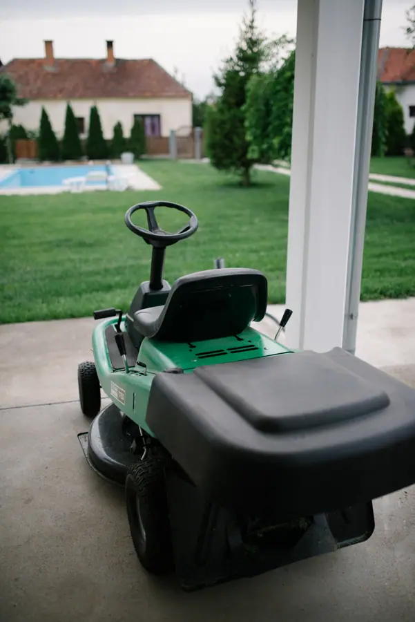 A lawn mower sits idle on a concrete patio, overlooking a well-kept garden and swimming pool
